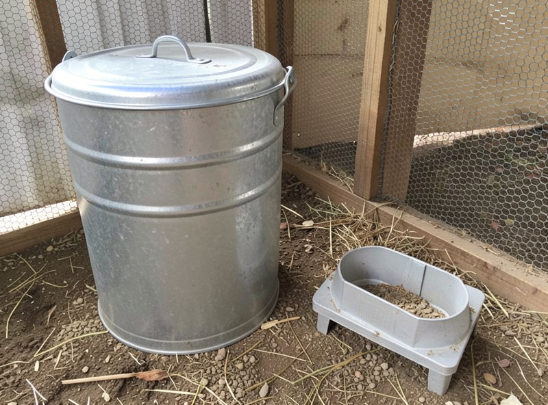 Galvanized steel feed storage can with sealed lid next to a treadle feeder in a coop