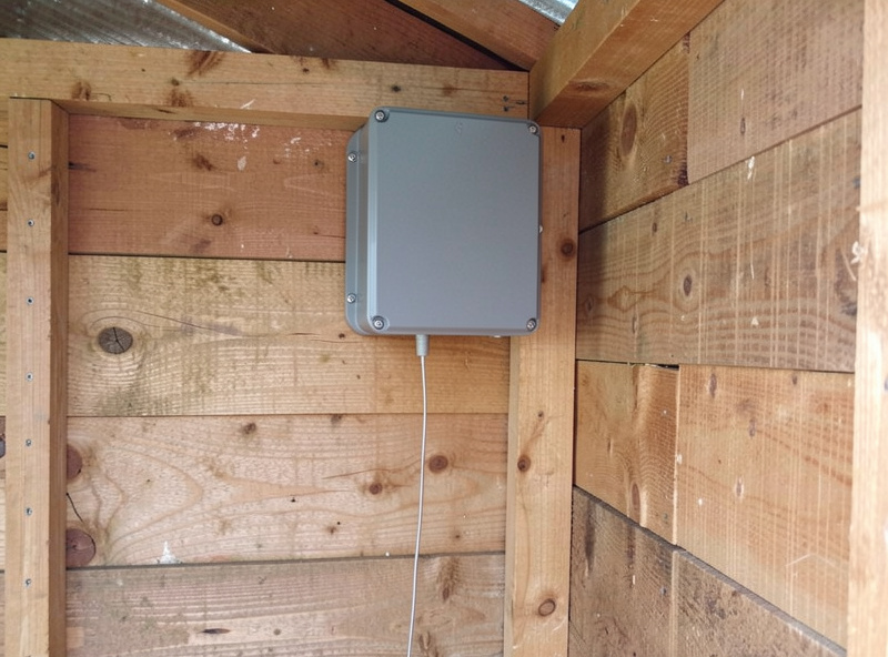 Small sealed electronics enclosure mounted high inside a wooden chicken coop with a sensor probe