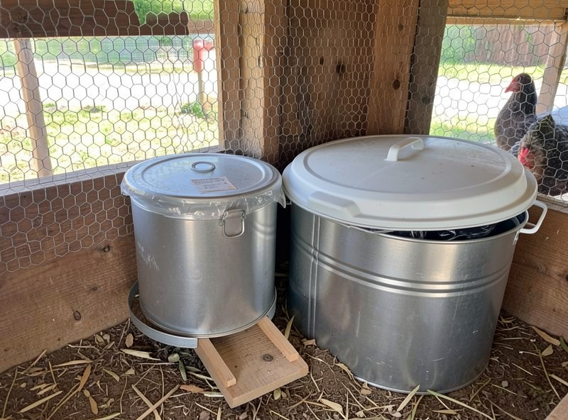 A treadle chicken feeder with its lid closed beside sealed steel feed cans in a coop