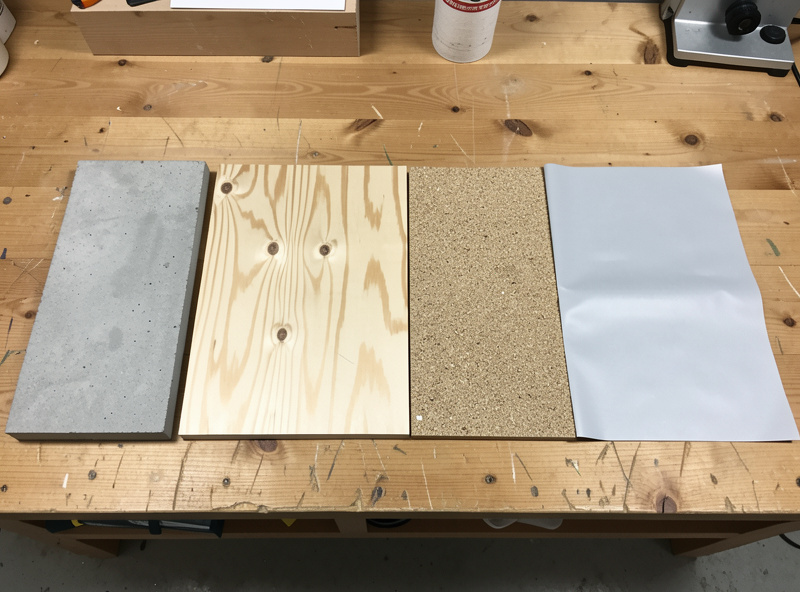 Comparison flat-lay of four coop flooring material samples — concrete, sealed plywood, coarse sand, and vinyl sheet — on a workshop bench